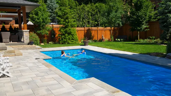 A backyard with hardscaping and a couple people swimming in a long pool