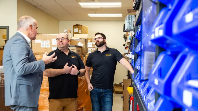A North Easton Savings Bank lender standing and talking to two local business partners in an storage room with various plastic part containers on shelves next to them