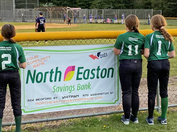 A few softball players with green t-shirts that say North Easton Savings Bank on the back standing next to a banner that has the North Easton Savings Bank logo on it as a sponsor