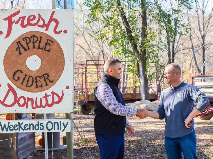 A North Easton Savings Bank lender shaking hands with a local business partner at a farm next to a hand-painted sign that says, "Fresh Apple Cider Donuts Weekends Only"